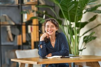 Portrait of smiling woman wearing spectacles and holding pencil while sitting at desk. Business woman taking notes in diary and looking at camera. University girl with eyeglasses sitting on table at library.