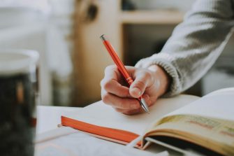 Close up of a person's hand holding an orange pen writing in an open notebook