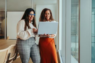 Two businesswomen working as a team in a tech startup environment. They are reviewing information on a laptop, reflecting a collaborative and innovative workplace atmosphere.