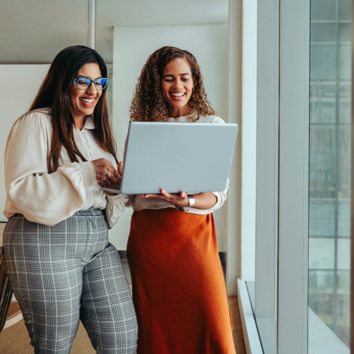 Two businesswomen working as a team in a tech startup environment. They are reviewing information on a laptop, reflecting a collaborative and innovative workplace atmosphere.