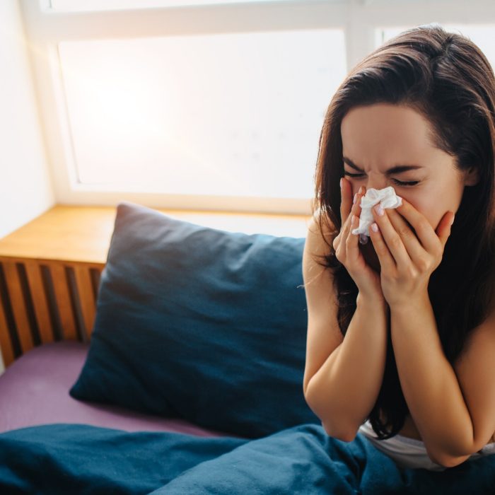 Young beautiful woman in morning bed at home. Blowing nose in white tisue. Flu or caught cold. Suffer from high temperature or virus