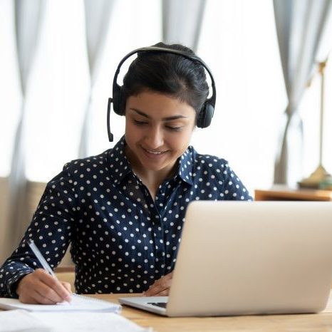 Happy indian girl student wear headset study online with webcam teacher write notes, happy young woman listen lecture watch webinar on laptop sit at desk, distance e learn language education concept