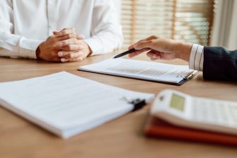 A business person indicates to a document as a man with interlocked hands observes