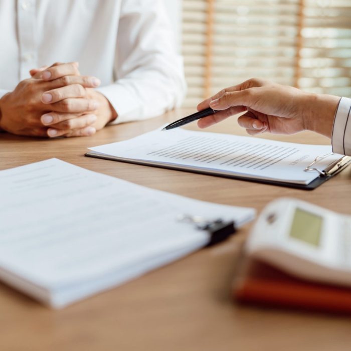 A business person indicates to a document as a man with interlocked hands observes