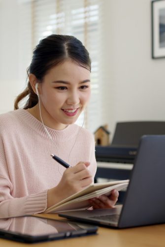 Candid young attractive asian female employee wear earphone listen to digital online course development program in quarantine social distance at home reskill upskill school class for career path.