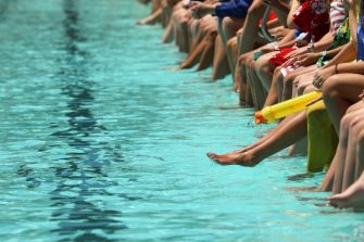 A bright aqua blue swimming pool with students sitting dangling their feet and toes in the water spectating. High school swimming carnival or club race meeting. 