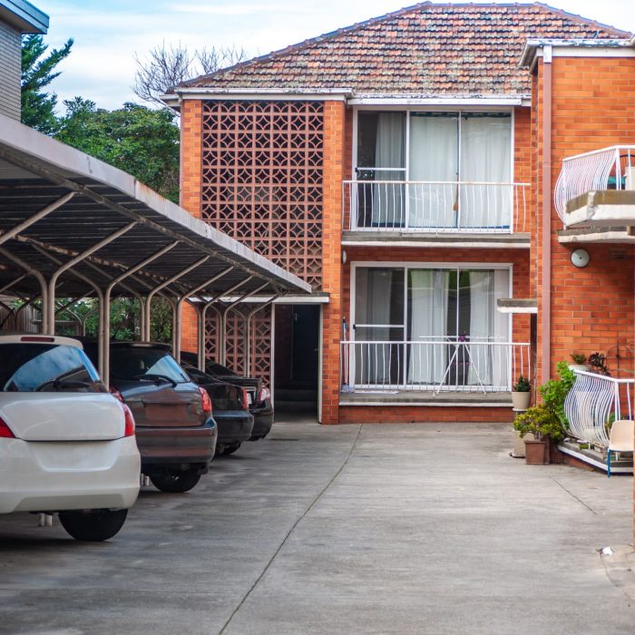 Covered parking area with parked cars sits adjacent to a two-story brick apartment building in an inner suburb of Australia.Residential complex features a mid-century architectural design with carport
